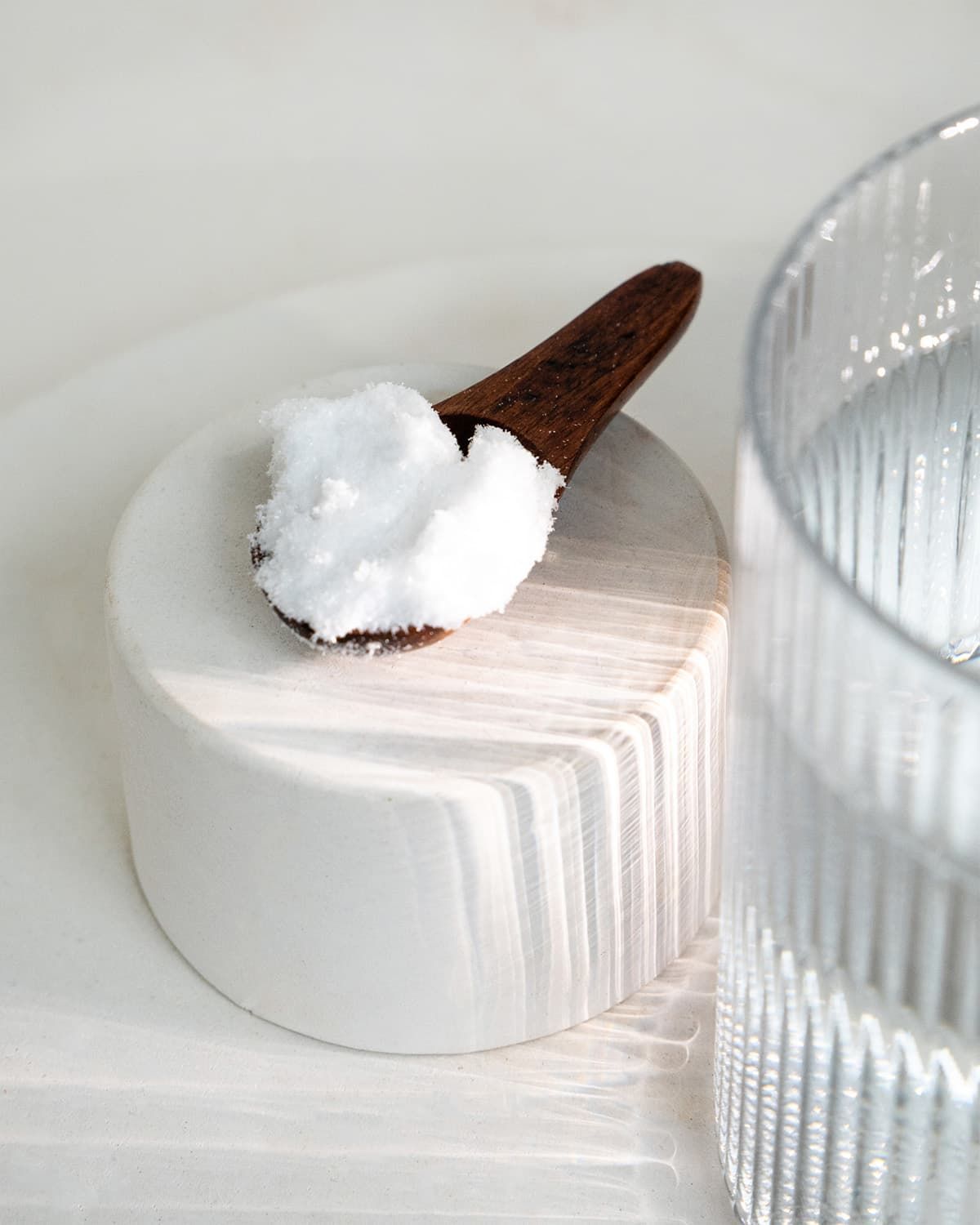 Ein Holzlöffel mit weißem Pulver auf einem weißen Zylinder, neben einem Glas Wasser.