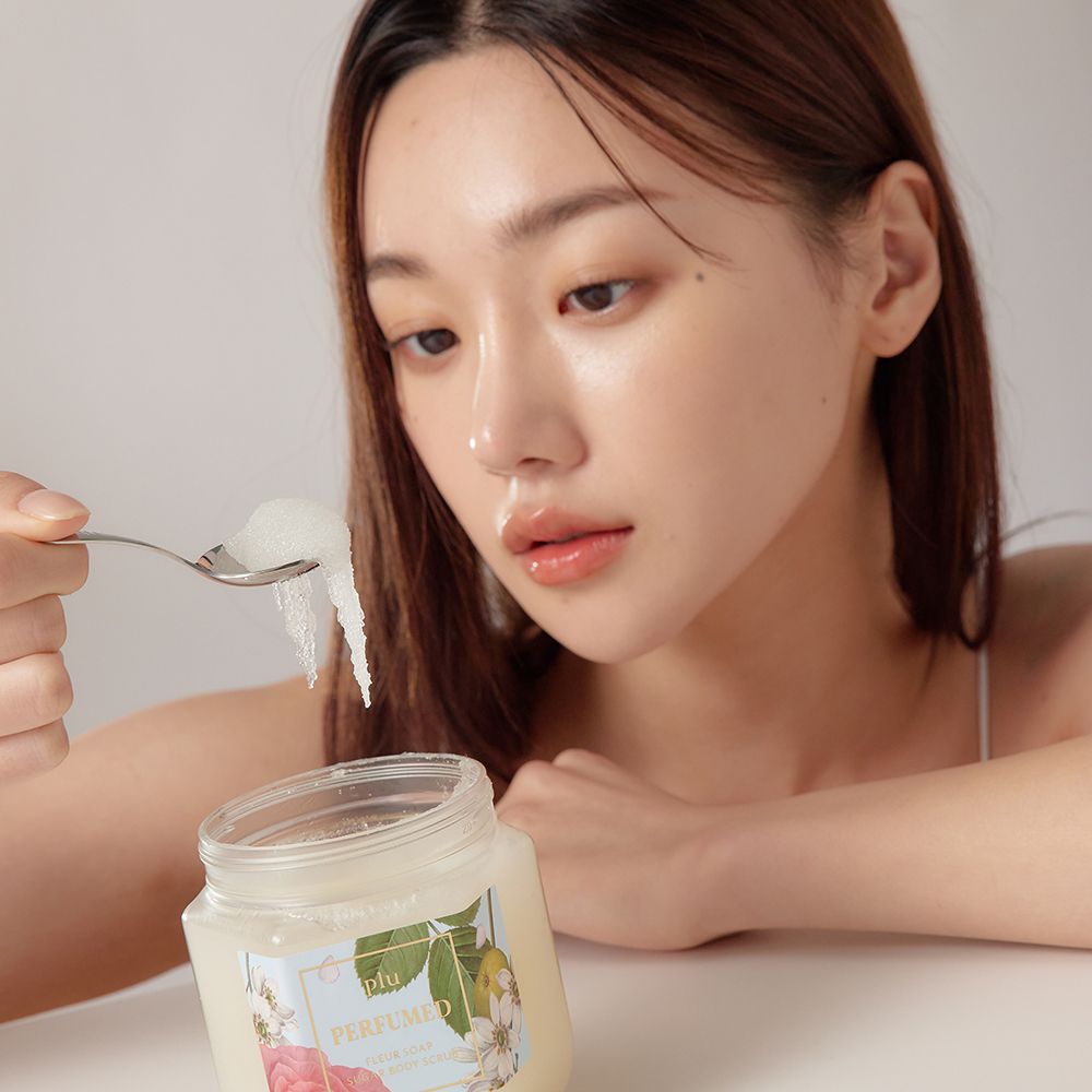 Person hält Löffel mit Körperpeeling über einem Glasbehälter. Aufschrift: PERFUMED FLEUR SOAP SUGAR BODY SCRUB.