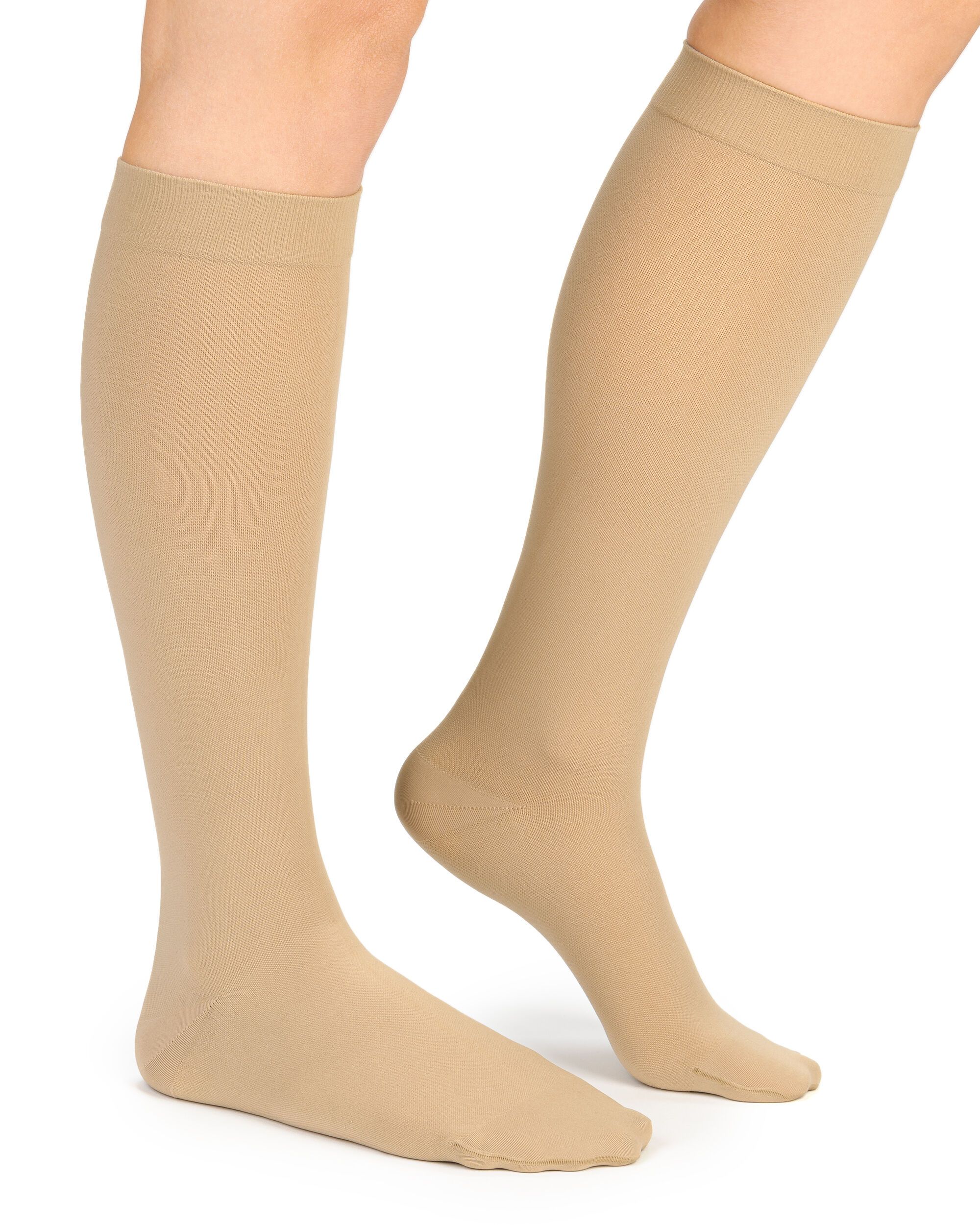 Beige Kompressions-Kniestrümpfe an Beinen. Blick von der Seite, Füße auf dem Boden. Glatter Stoff, ohne Muster.