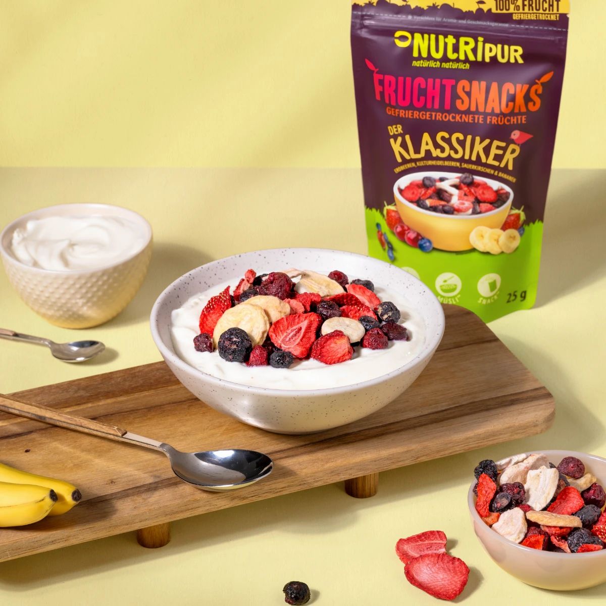 Schale mit Früchten auf Joghurt. Daneben Beutel mit gefriergetrockneten Früchten. Löffel und Schale mit Joghurt.