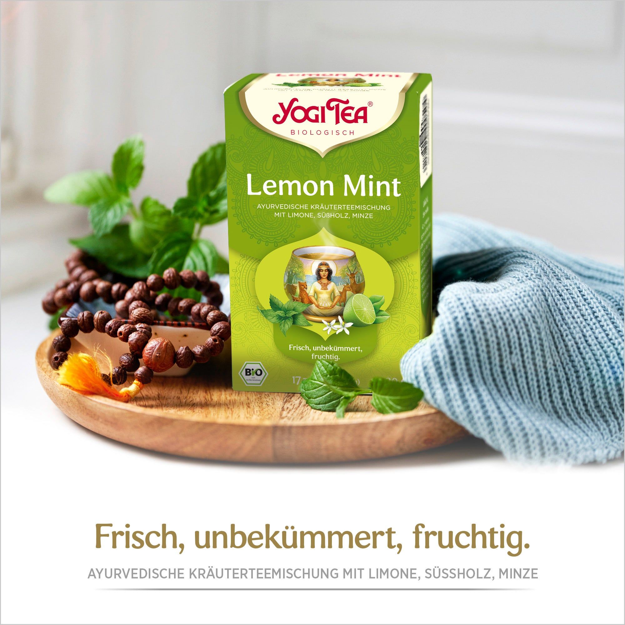 YOGI TEA® Lemon Mint-Packung auf Holztablett, umgeben von Minze und Perlenkette. Text: Frisch, unbekümmert, fruchtig.