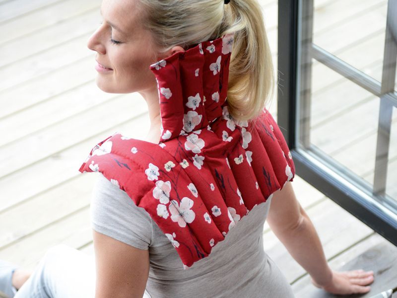 Person mit roter, floral gemusterter Leinsamenkompresse am Nacken. Frau sitzt am Fenster.