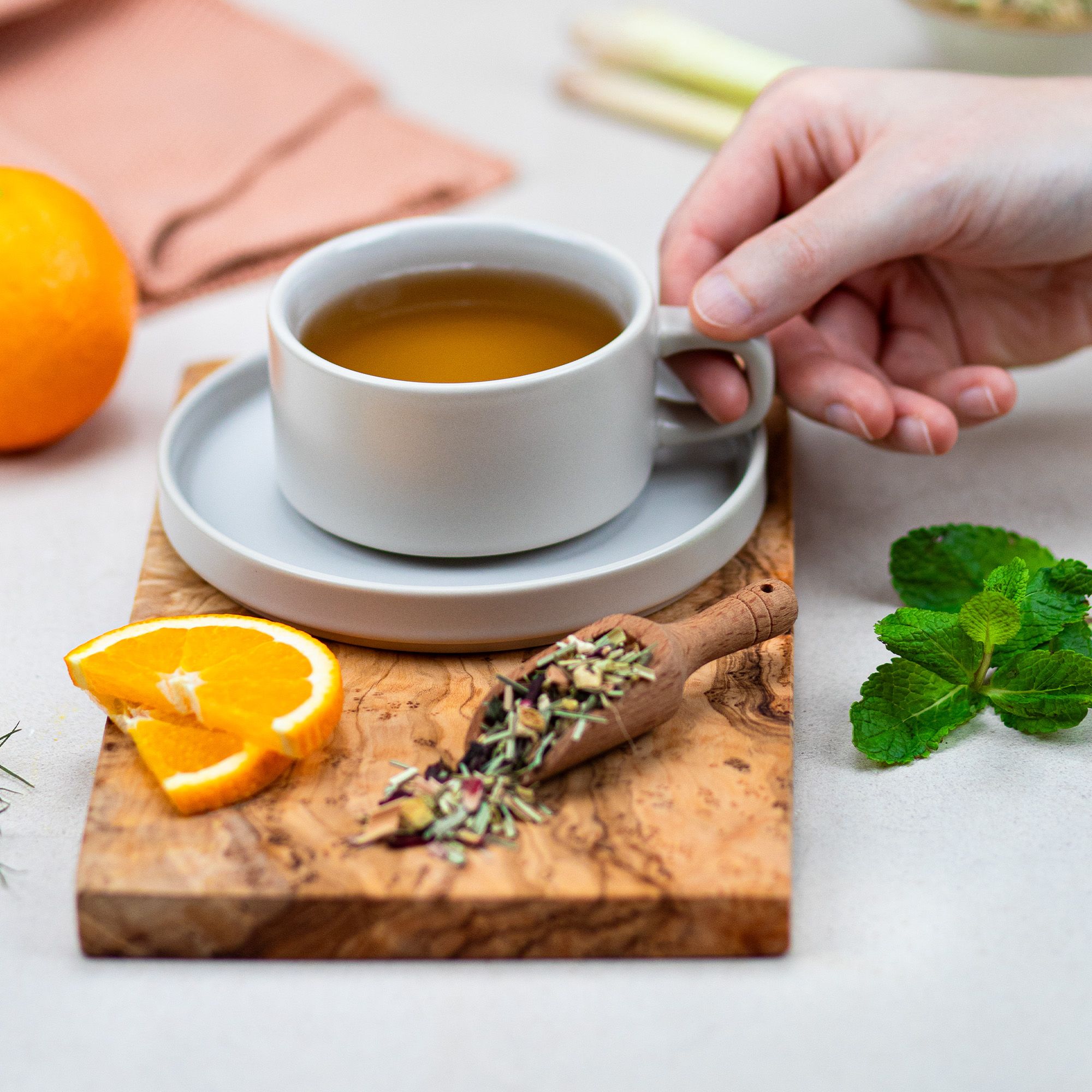 Tasse Tee auf Untertasse, Holzbrett, Orangenscheiben, Minze, Holzlöffel mit Kräutern. Hand hält Tasse.