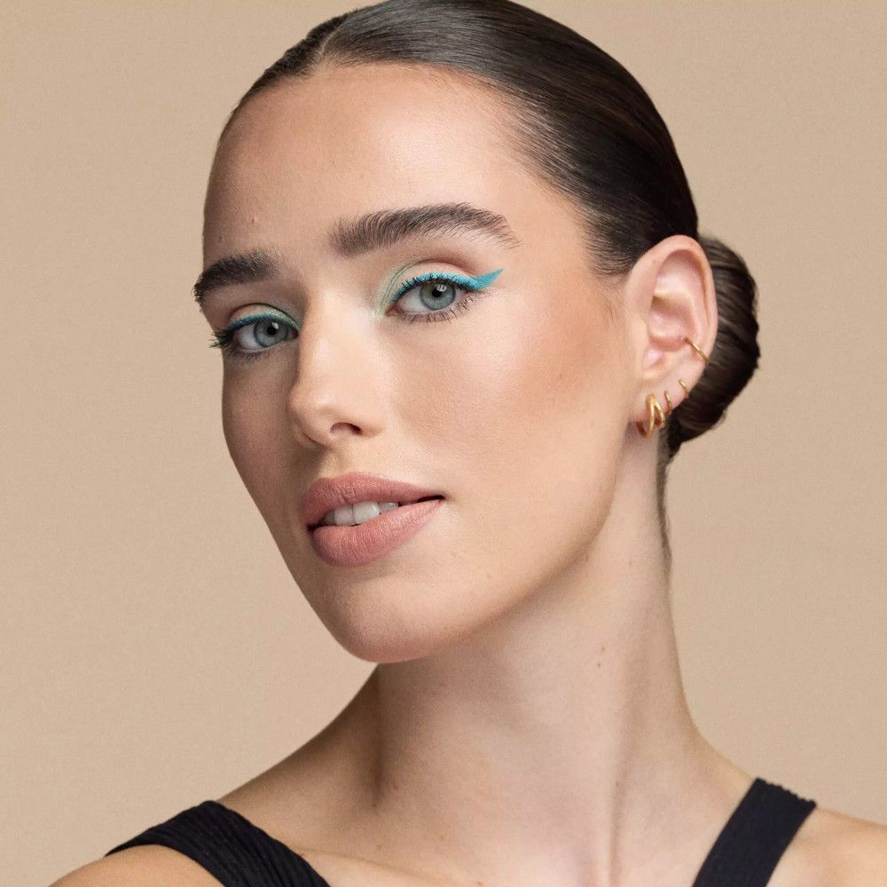 Porträt einer Frau mit blauem Eyeliner und rosafarbenem Lippenstift. Sie hat einen Dutt und trägt ein schwarzes Top.