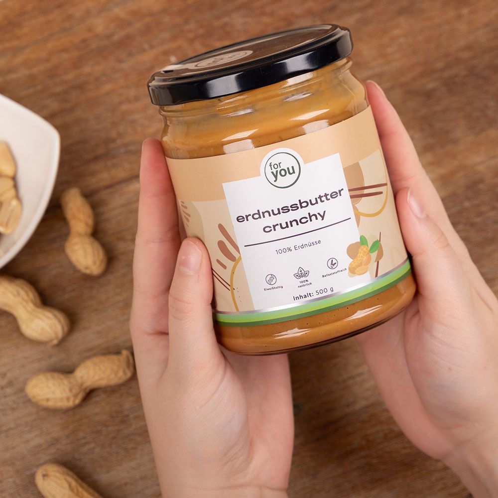 Glas Erdnussbutter Crunchy in Händen. Aufschrift: for you, Erdnussbutter Crunchy, 100% Erdnüsse, Inhalt: 500 g. Daneben Erdnüsse.