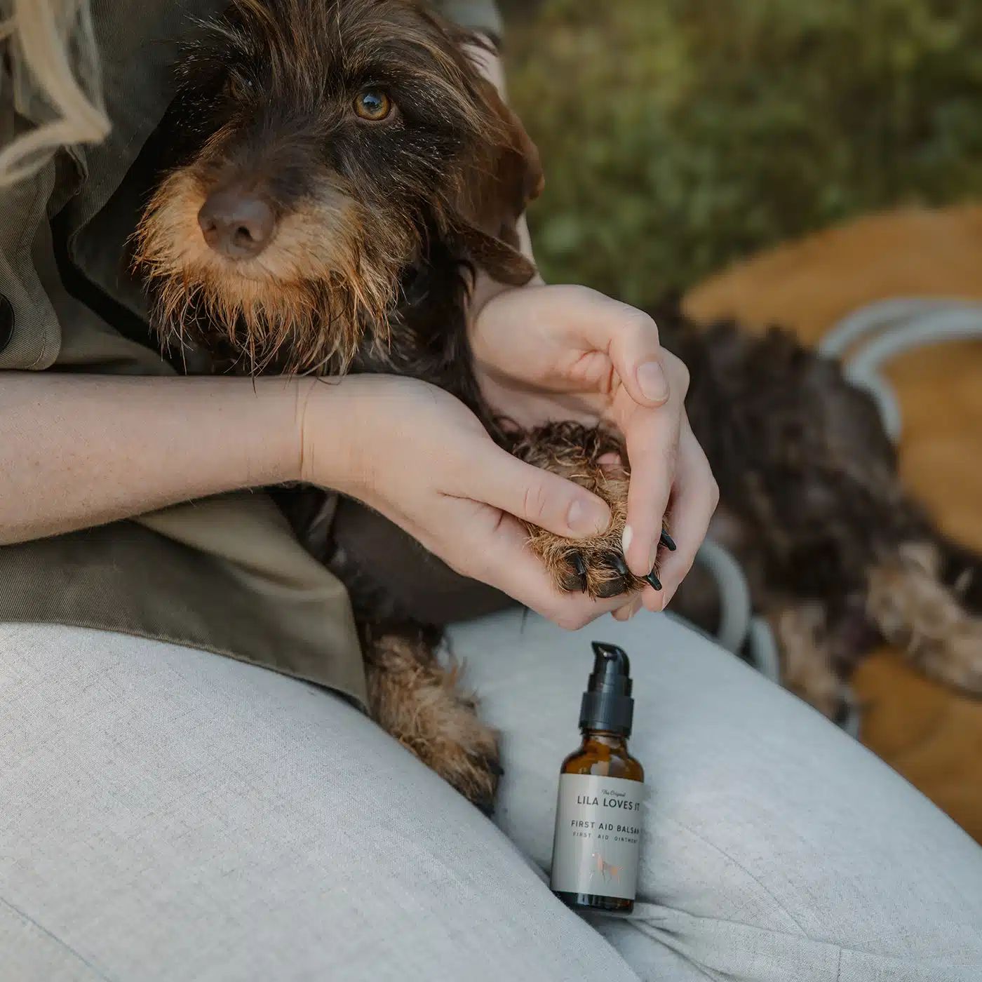 Person hält Hundepfote. Daneben steht eine braune Flasche mit schwarzem Pumpkopf. Aufschrift: LILA LOVES IT, FIRST AID BALSAM.