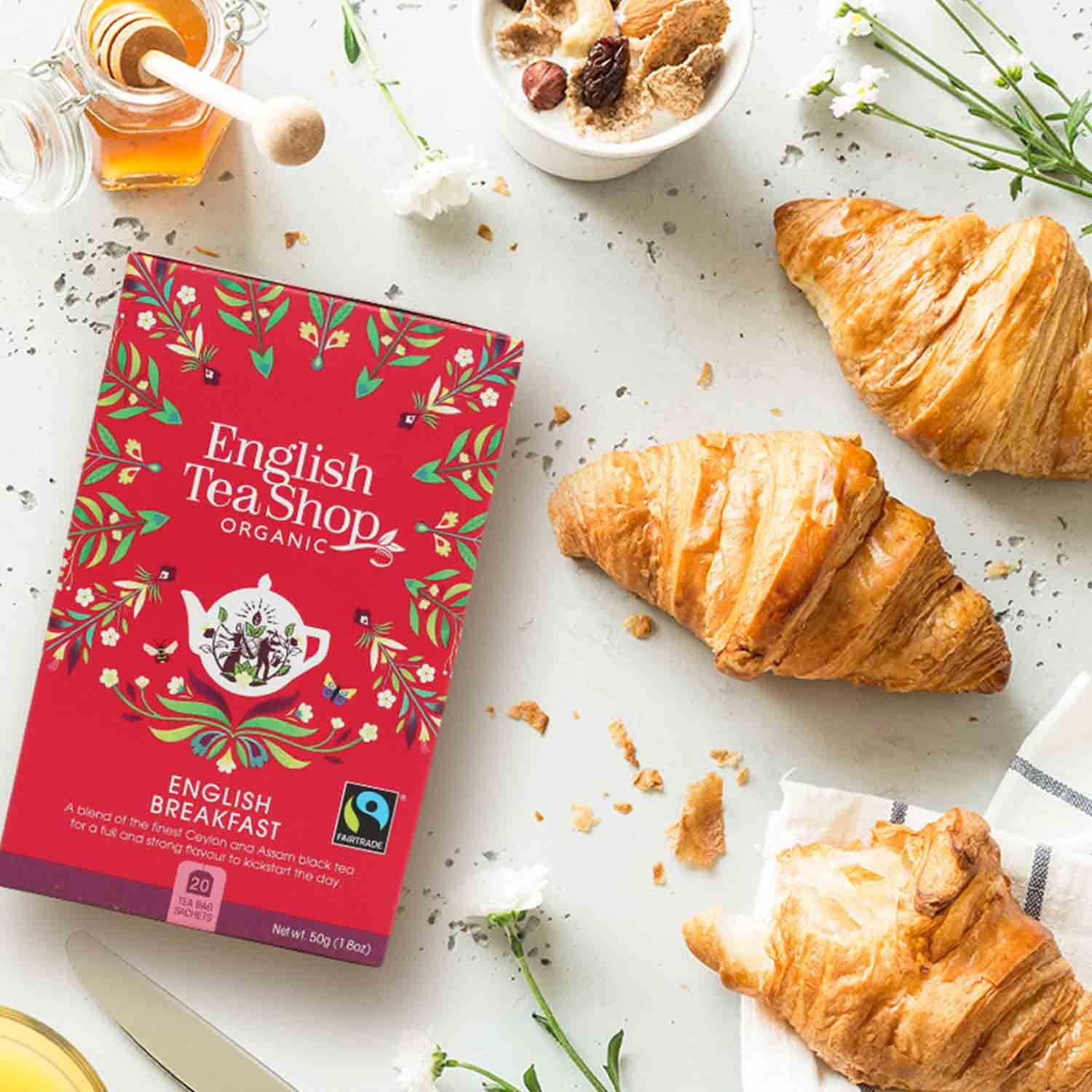 Rote "English Tea Shop Organic"-Verpackung neben Croissants, Honig, Müsli und Blumen auf hellem Untergrund.