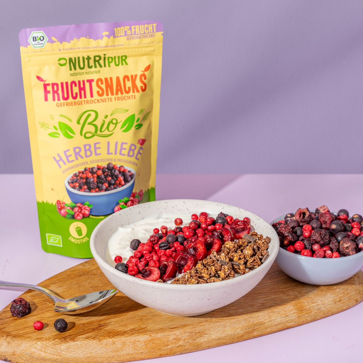 Produktverpackung und Schalen mit gefriergetrockneten Früchten und Joghurt auf Holzbrett. Löffel daneben. Bio-Siegel.