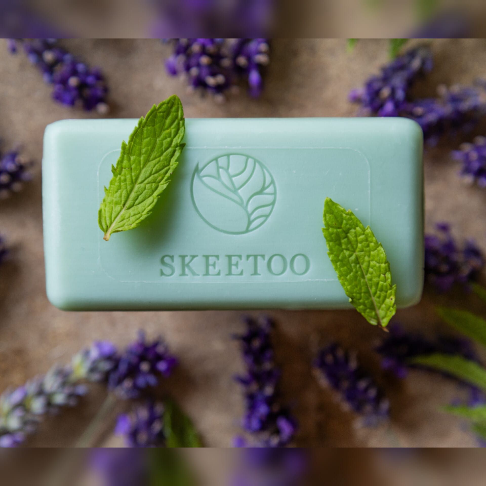 Hellgrünes Seifenstück mit Logo und Schriftzug SKEETOO. Zwei grüne Blätter liegen auf der Seife. Lavendelblüten umgeben die Seife.