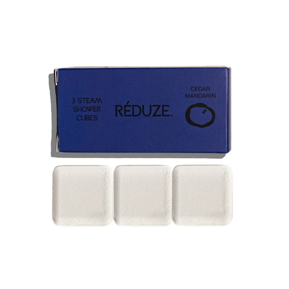 Drei weiße Duschwürfel und eine blaue Schachtel mit Produktinformationen. RÉDUZE Duschwürfel Zedernholz & Mandarine.