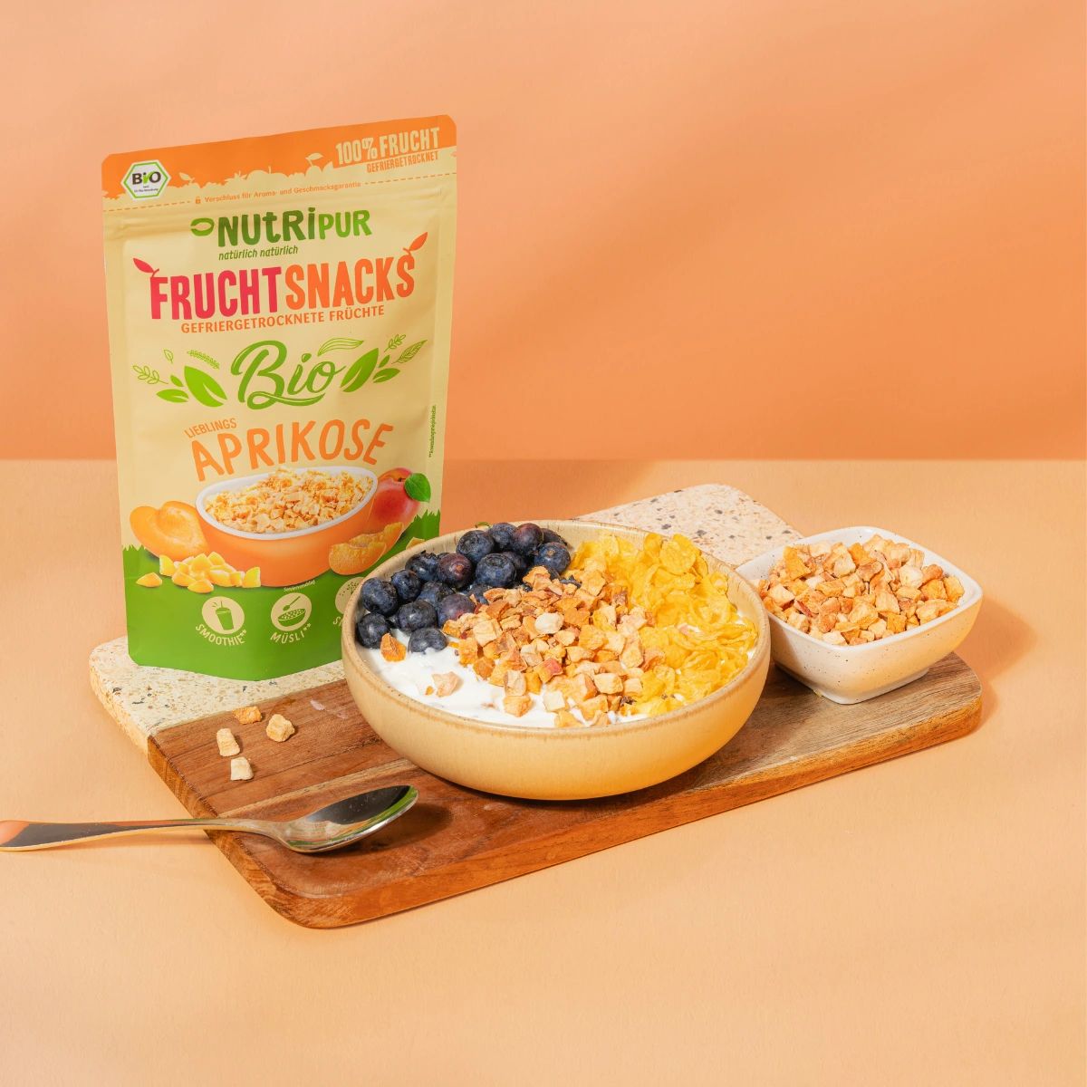 Schale mit Joghurt, Blaubeeren und gefriergetrockneten Aprikosen. Verpackung NutriPur Fruchtsnacks. Auf Holzbrett.