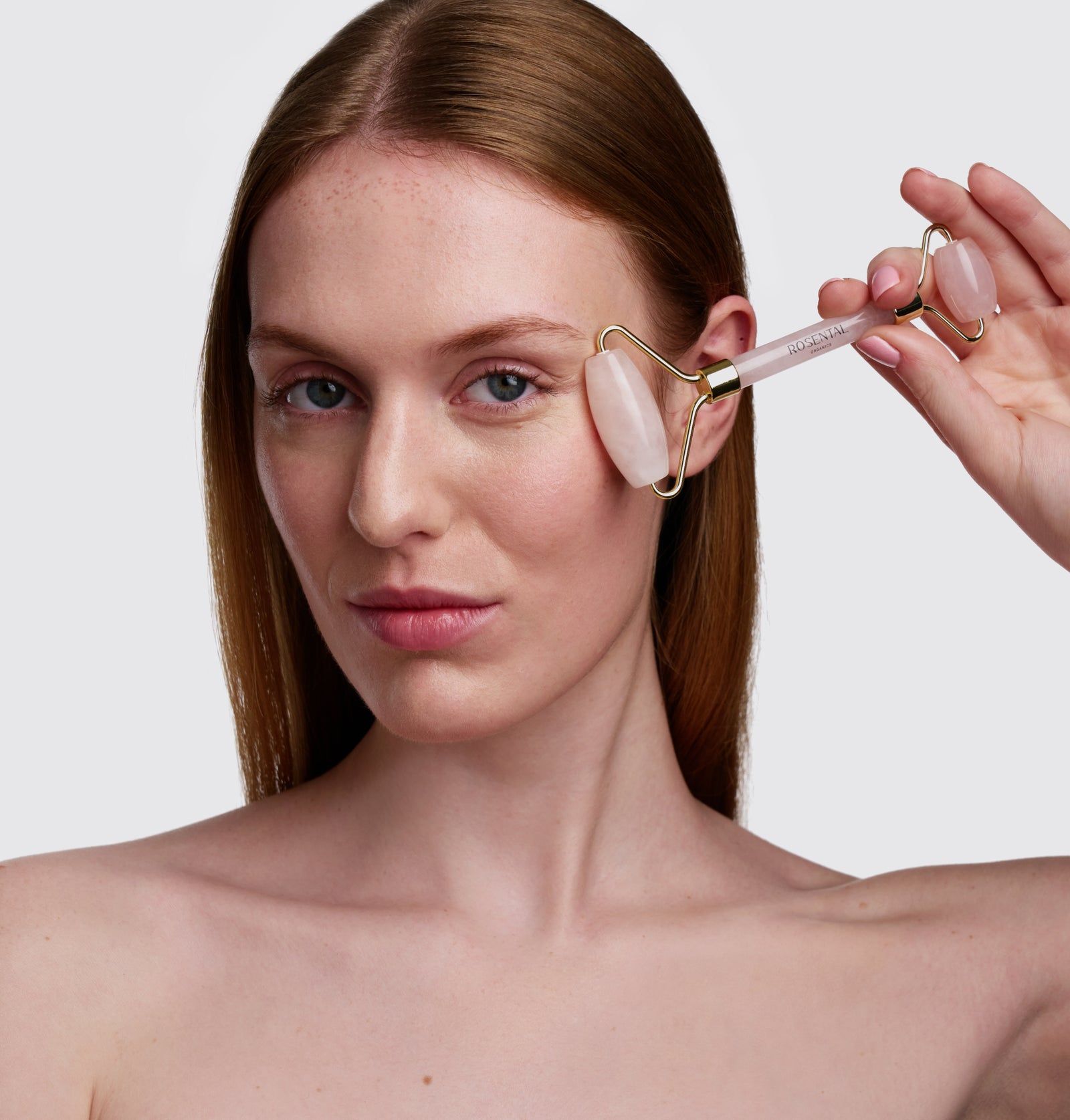 Frau mit Rose Quartz Roller am Auge. Goldfarbener Rahmen, rosa Rollen. Produktname nicht sichtbar.