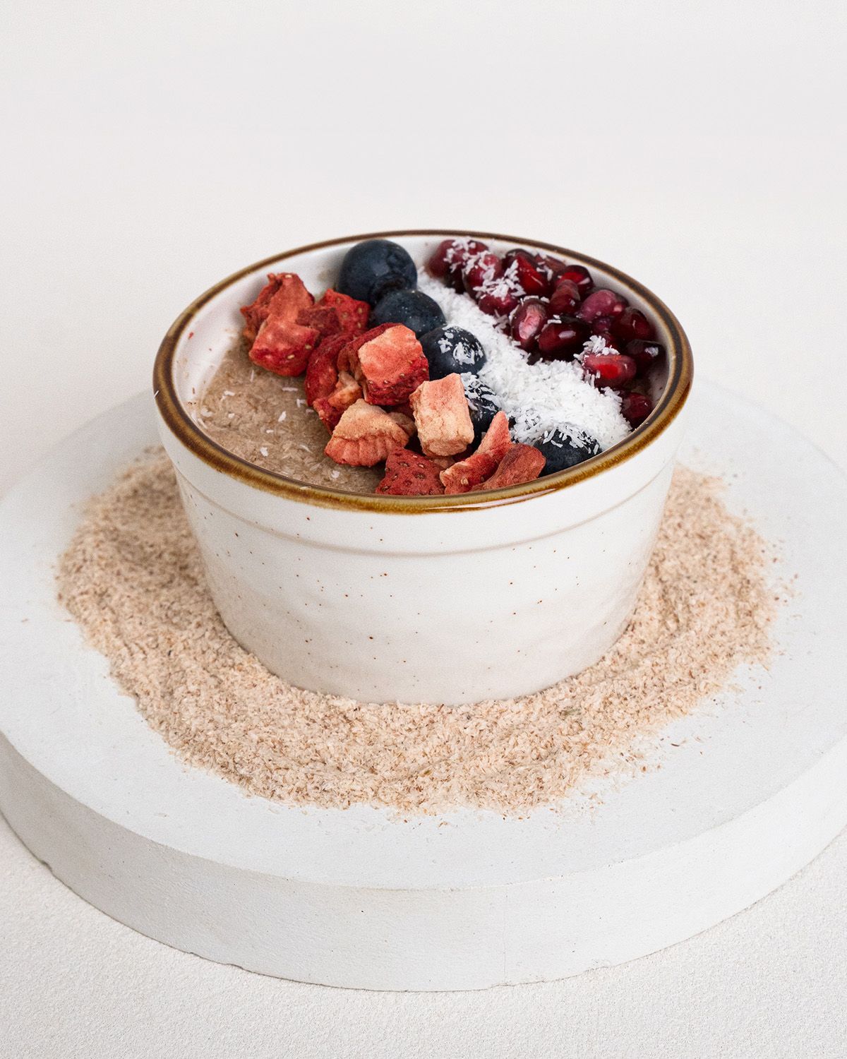 Schale mit Getreidebrei, belegt mit Beeren und Kokosraspeln. Auf einer Unterlage aus braunen Schalen. Auf weißem Hintergrund.