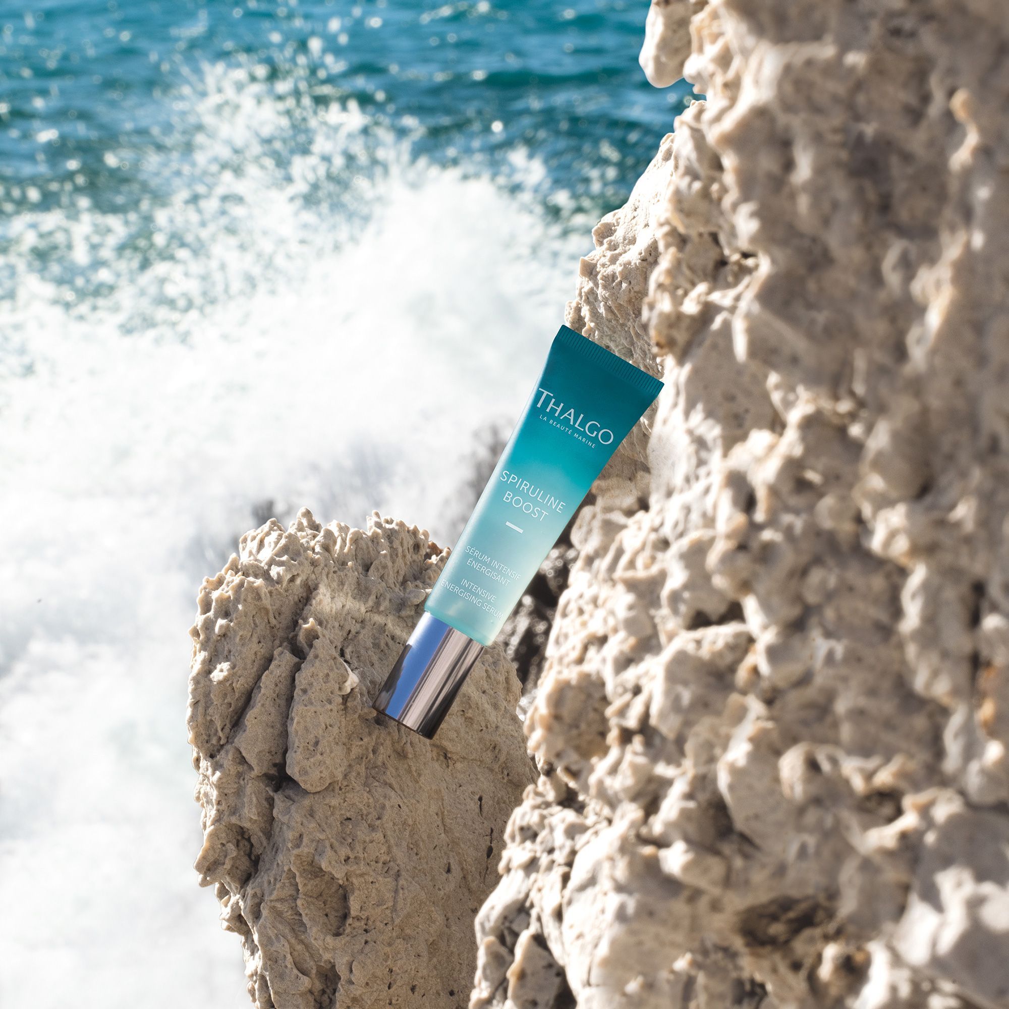 THALGO SPIRULINE BOOST Serum-Tube auf Felsen am Meer. Türkis-weißes Design. Silberfarbener Pumpkopf. Wellen im Hintergrund.