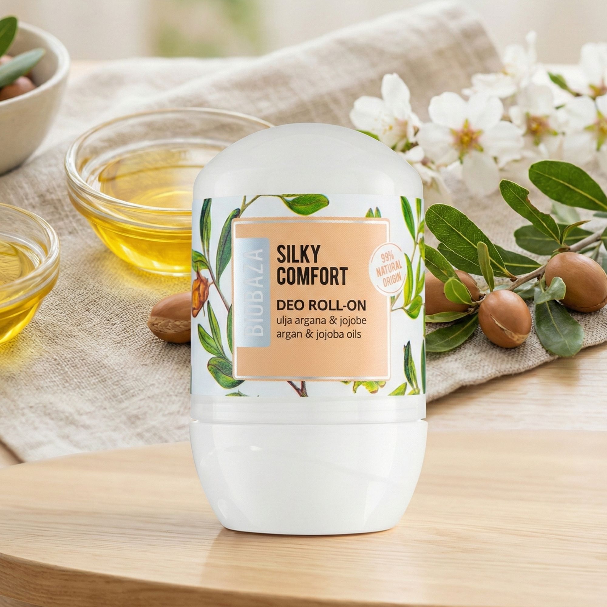Weißer Deo Roll-on mit Etikett. Aufschrift: Silky Comfort, Deo Roll-on, Argan- und Jojobaöle. Hintergrund: Arganfrüchte, Öl und Blüten.