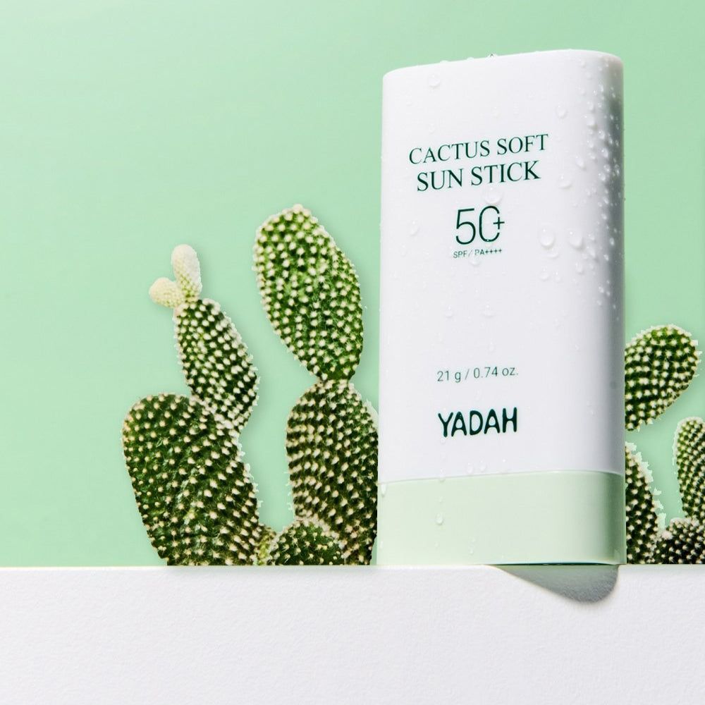 Ein weißer Sonnenschutzstift mit grüner Unterseite vor einem Kaktus. Auf dem Stift steht 'CACTUS SOFT SUN STICK 50' und 'YADAH'.