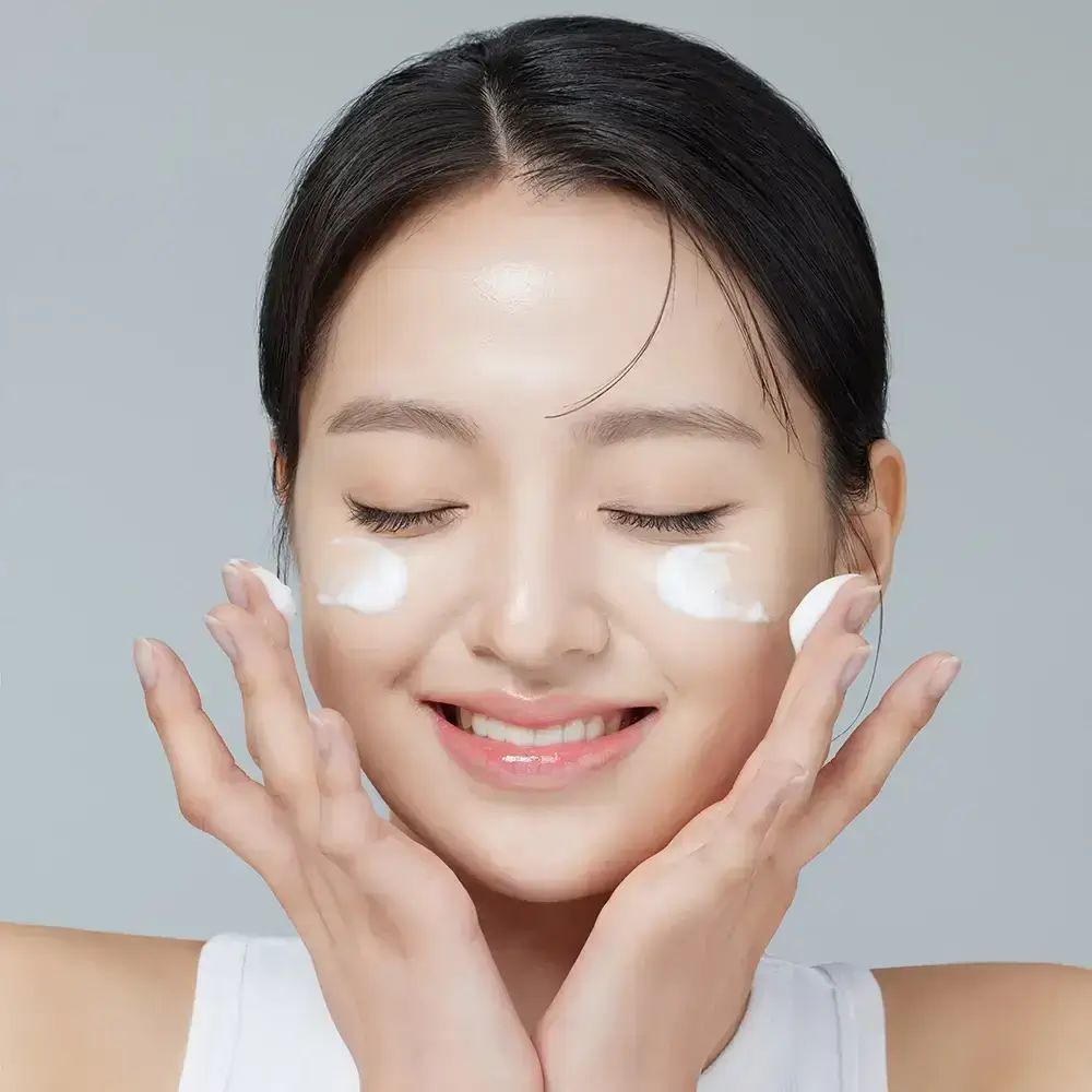 Frau mit geschlossenen Augen, die Creme auf ihr Gesicht aufträgt. Sie lächelt. Weißes Top.