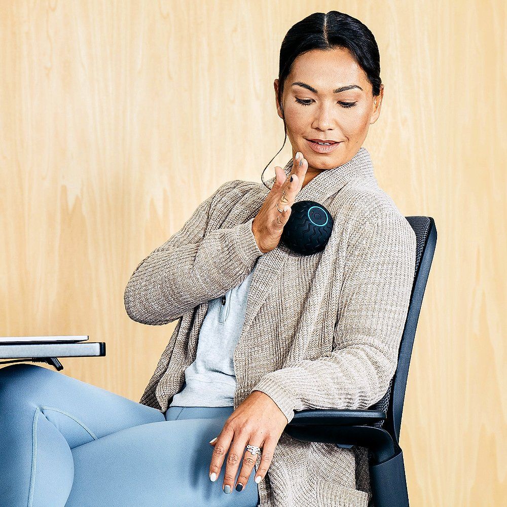 Frau sitzt und hält den schwarzen Massageball an der Schulter. Sie trägt eine Strickjacke und blaue Hose. Holz-Hintergrund.
