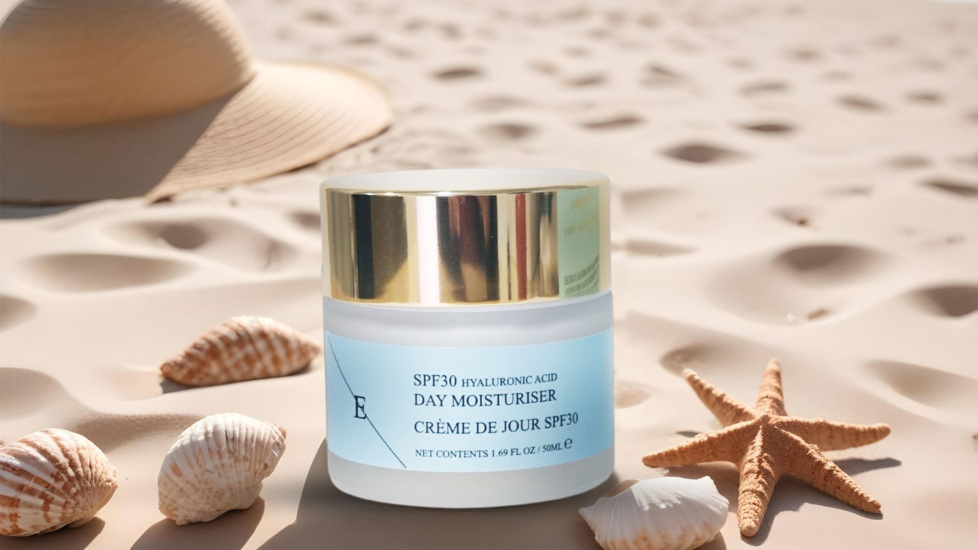 Ein Cremetiegel mit goldenem Deckel und blauem Etikett. Auf Sand, mit Muscheln, Seestern und Hut im Hintergrund.