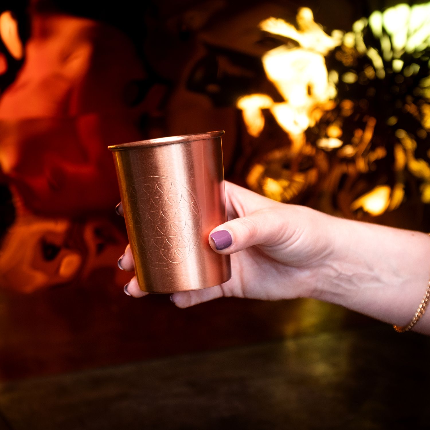 Hand hält Kupferbecher mit Blume des Lebens. Kupferfarben, glänzend. Hintergrund unscharf.