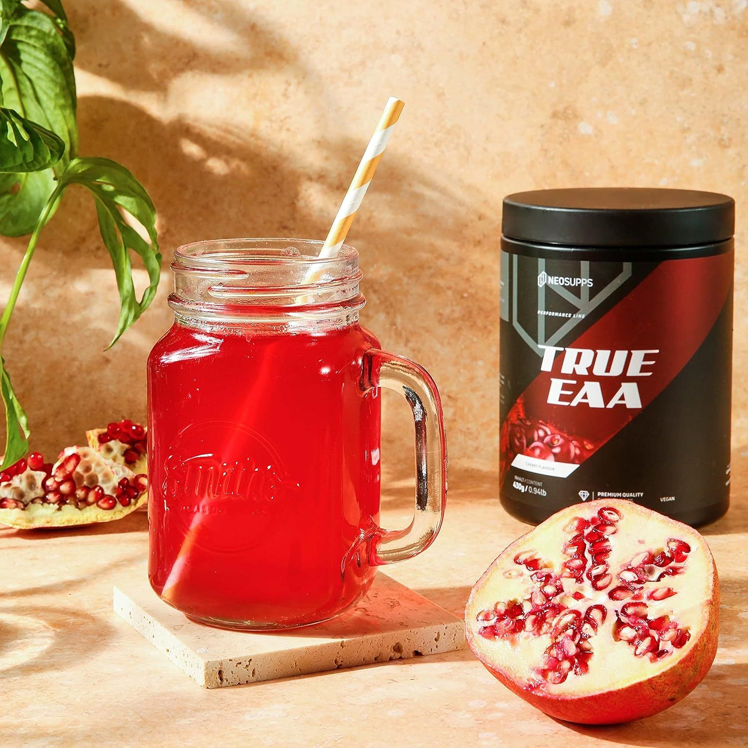 Glas mit rotem Getränk, Strohhalm. Daneben Dose TRUE EAA. Granatapfelhälfte. Aufschrift: TRUE EAA. 430g/0.94lb. Mit Granatapfel und Glas.