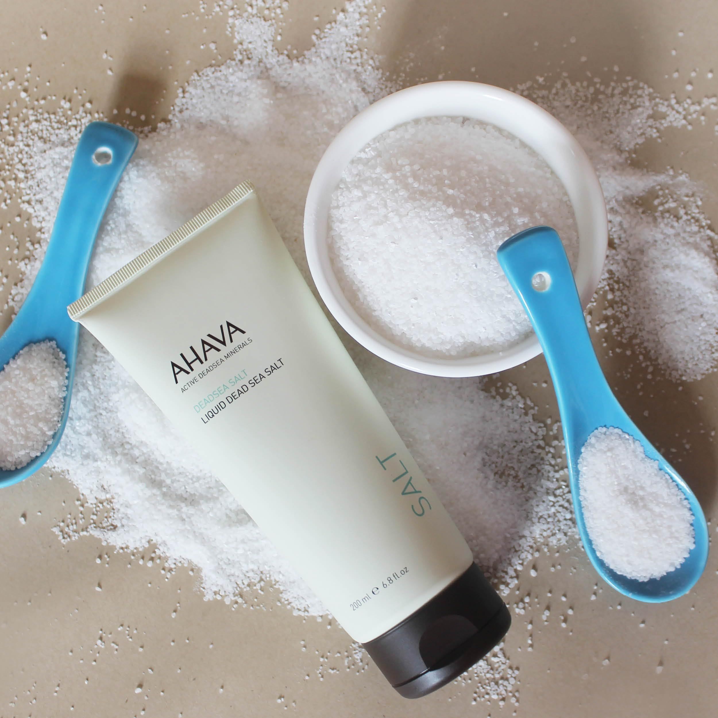 Beige Tube mit AHAVA-Logo und Text, umgeben von Salz und blauen Löffeln mit Salz. Eine Schale mit Salz ist ebenfalls vorhanden.