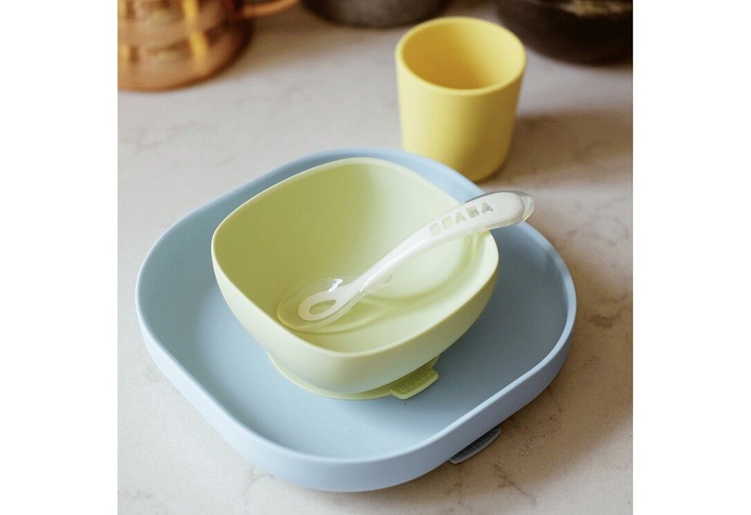 Geschirrset mit Teller, Schüssel, Becher und Löffel. Schüssel mit Logo. Farben: Blau, Grün und Gelb. Auf Tisch platziert.