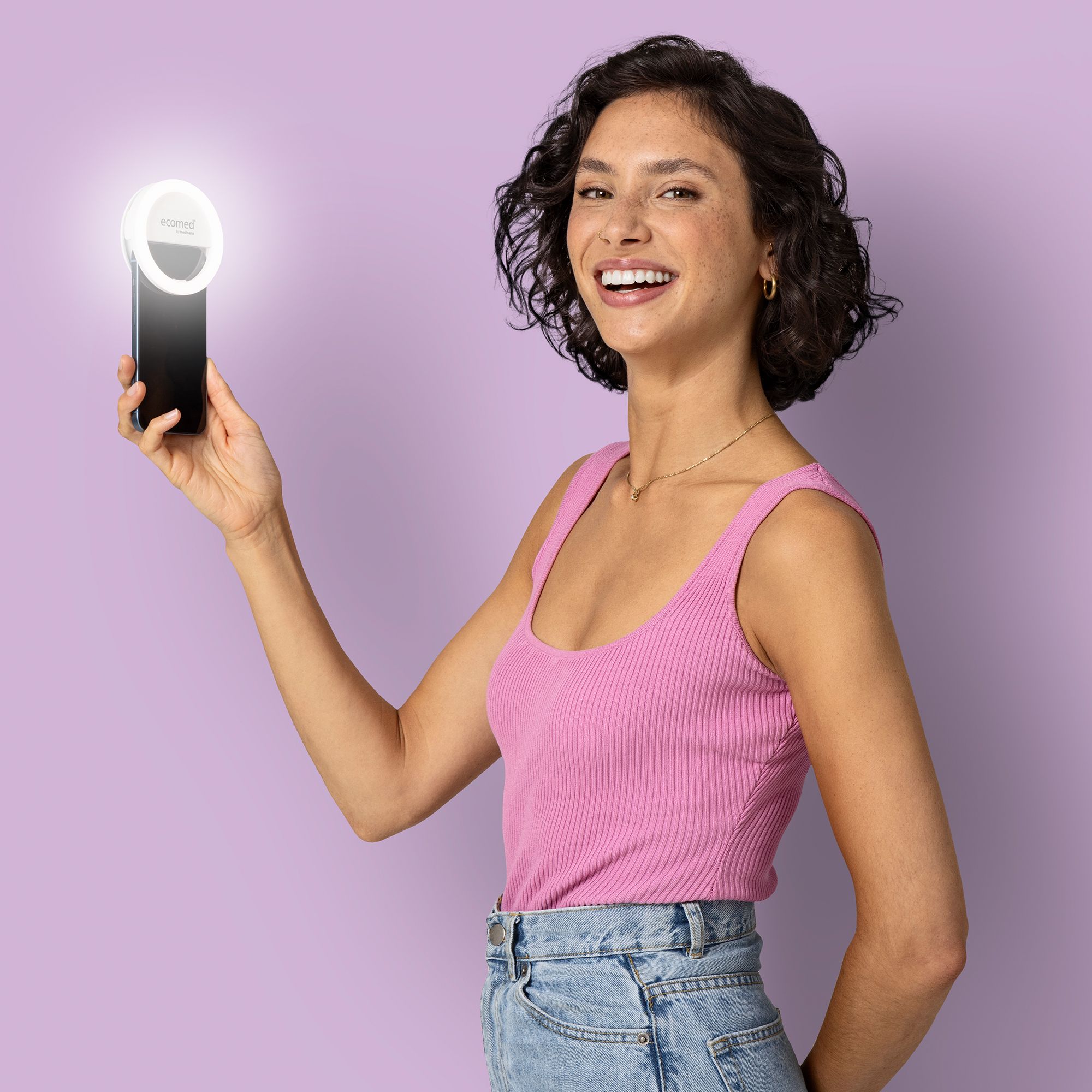 Eine Frau hält einen weißen Selfie-Spiegel mit LED-Beleuchtung vor einem Smartphone. Sie lächelt.