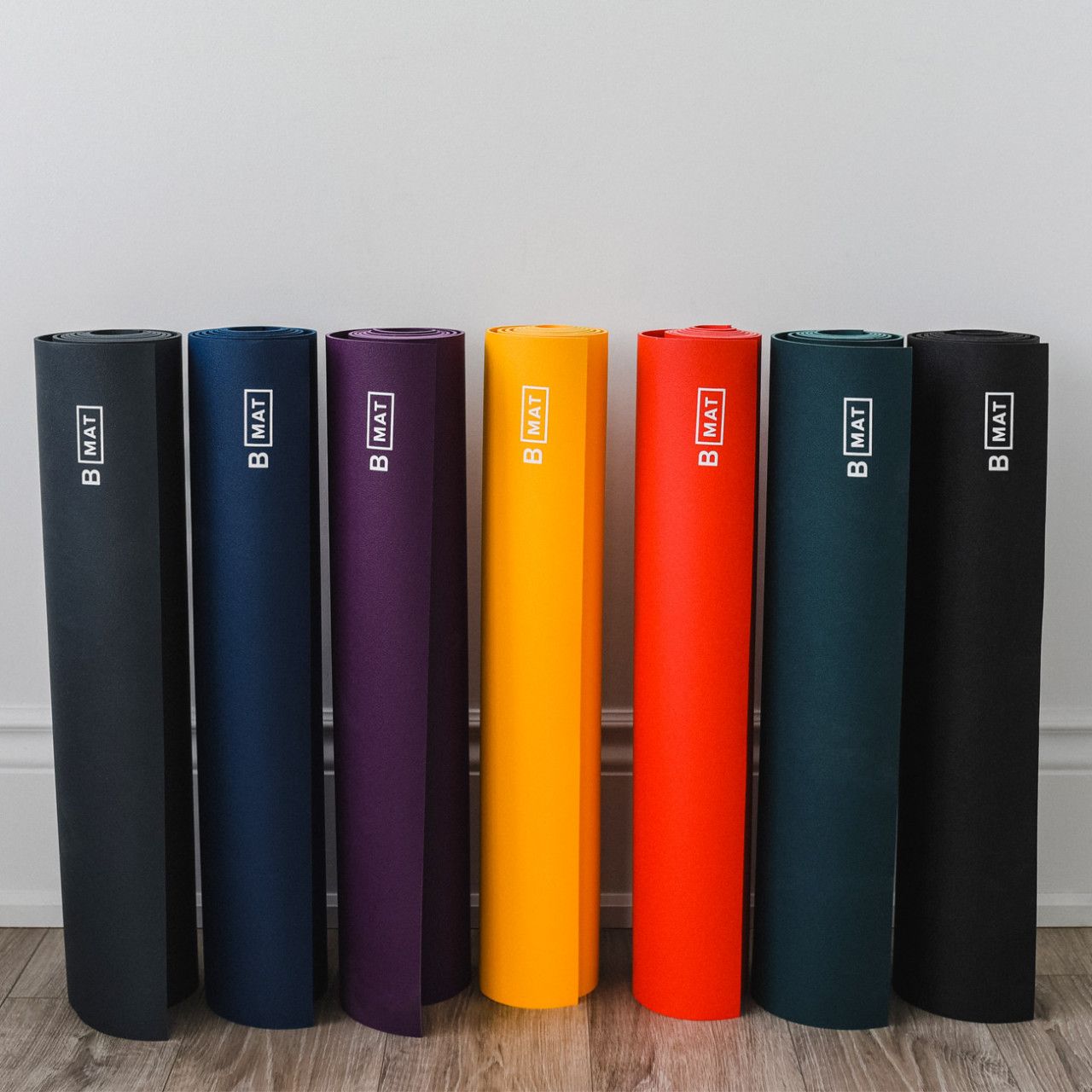 Sieben gerollte Yogamatten in verschiedenen Farben. Logo "B MAT" auf jeder Matte. Auf hellem Boden.