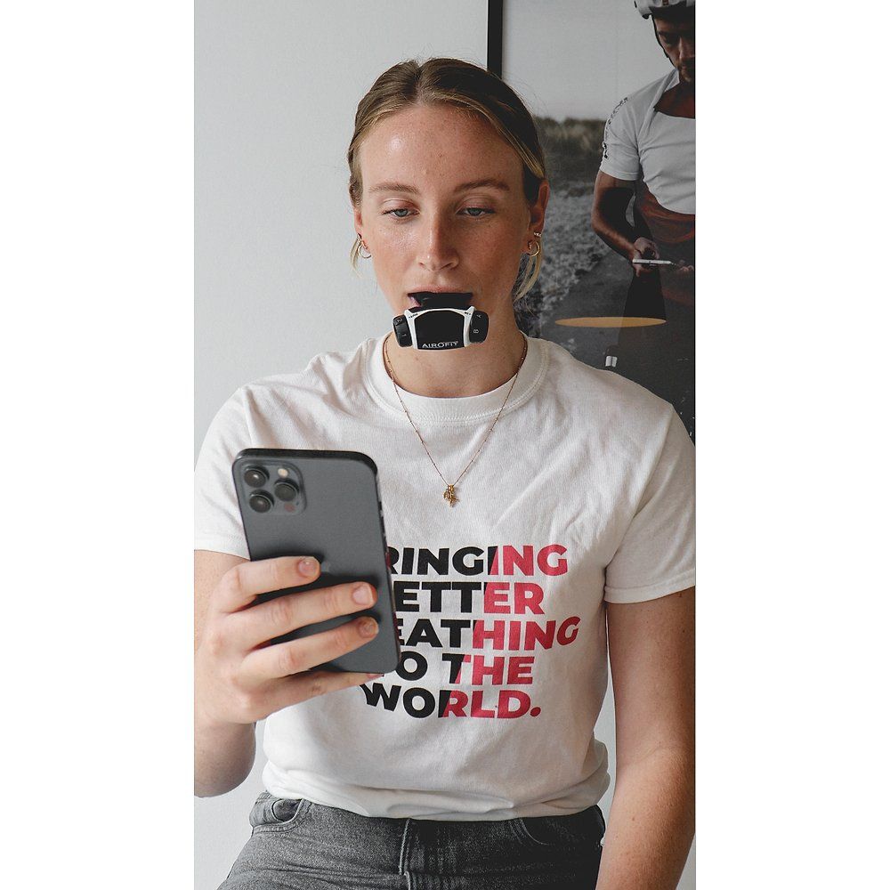 Frau mit Atemtrainer im Mund und Smartphone. Auf dem T-Shirt steht: BRINGING BETTER BREATHING TO THE WORLD.