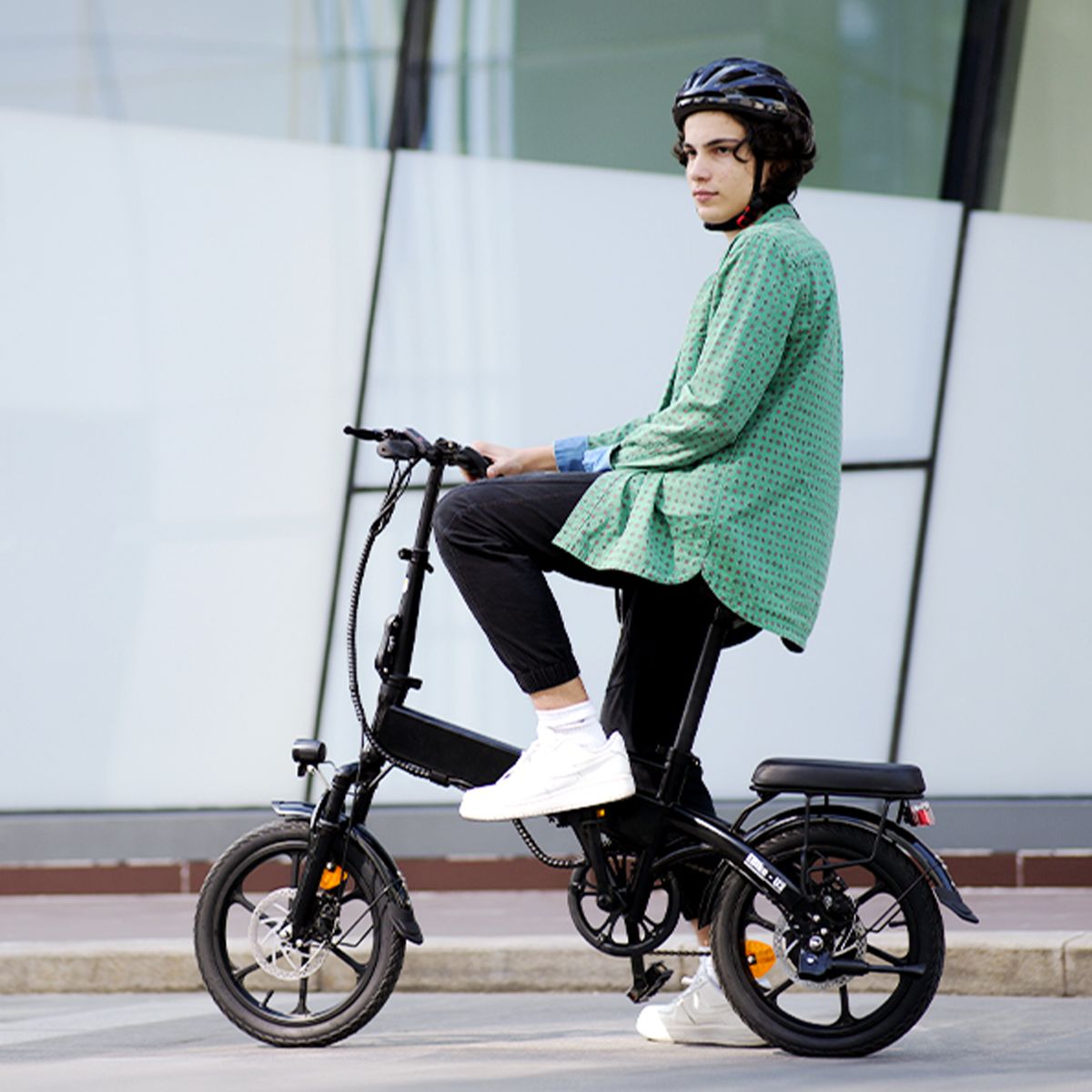Person sitzt auf einem schwarzen, zusammenklappbaren Elektrofahrrad. Trägt Helm und grüne Jacke. Hintergrund: Gebäude.