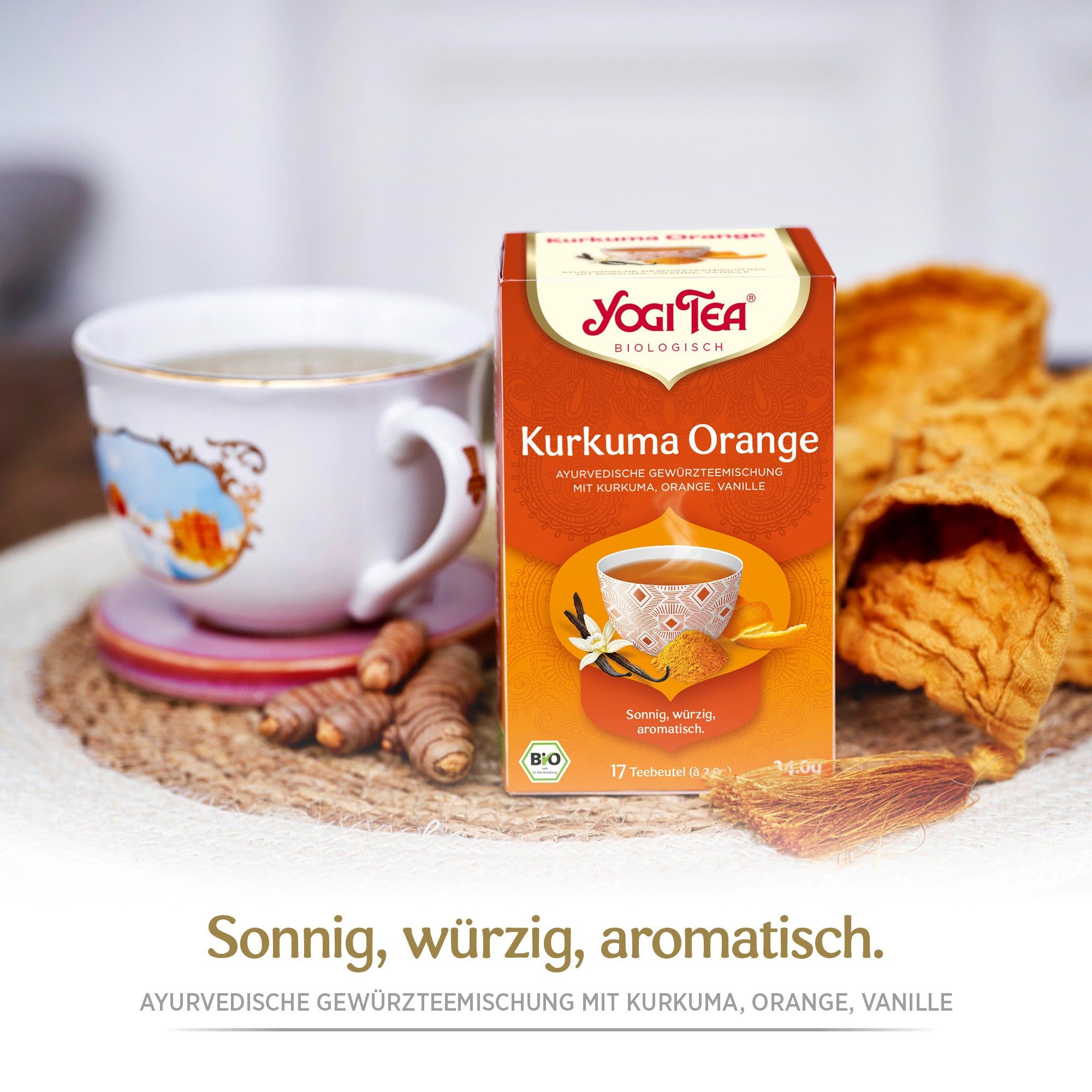 Yogi Tea Kurkuma Orange Bio Gewürztee Packung neben einer Tasse Tee und Kurkuma-Stücken. Text: Sonnig, würzig, aromatisch. Bio-Siegel.