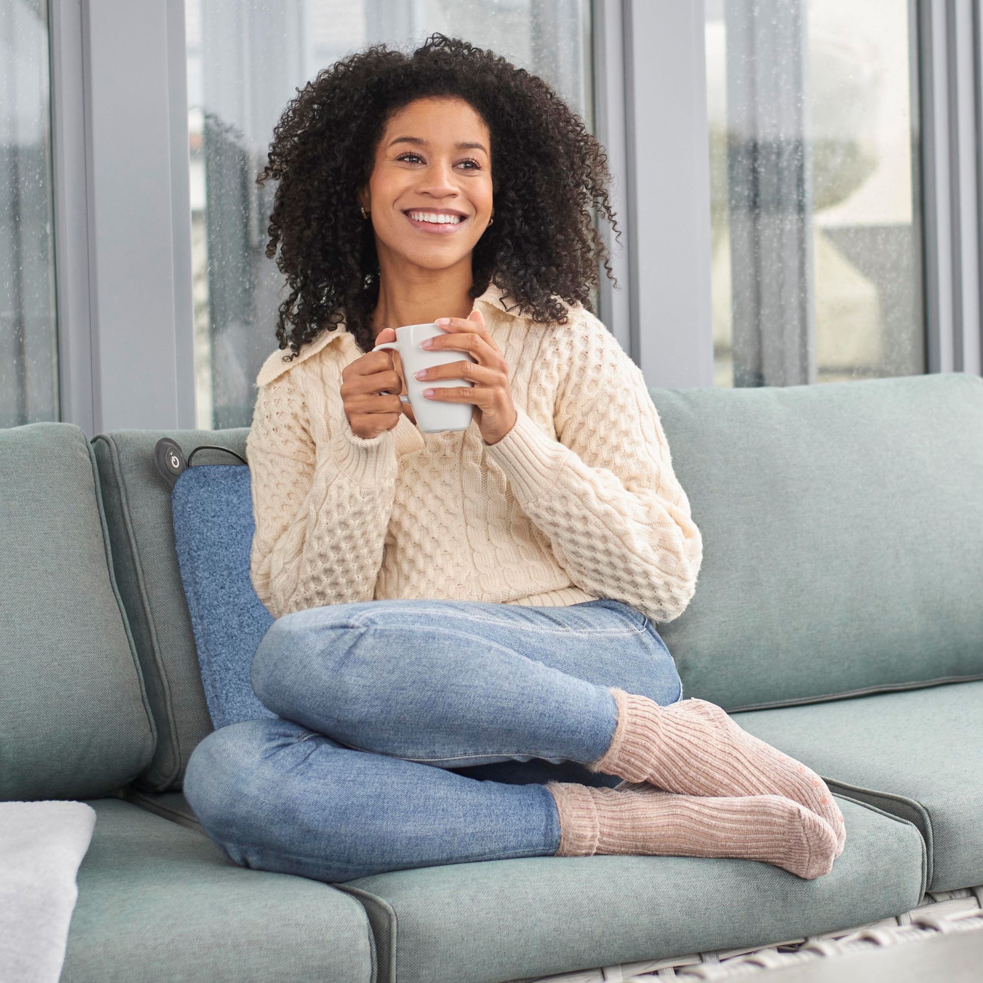 Frau sitzt entspannt auf einem Sofa mit einer Tasse in der Hand, trägt einen Pullover und Jeans.