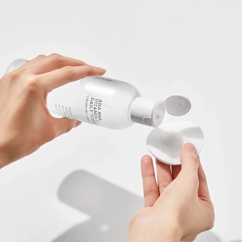 Weiße Flasche mit Toner, wird auf ein Wattepad gegossen. Aufschrift: AHA BHA Vitamin C Daily Toner.