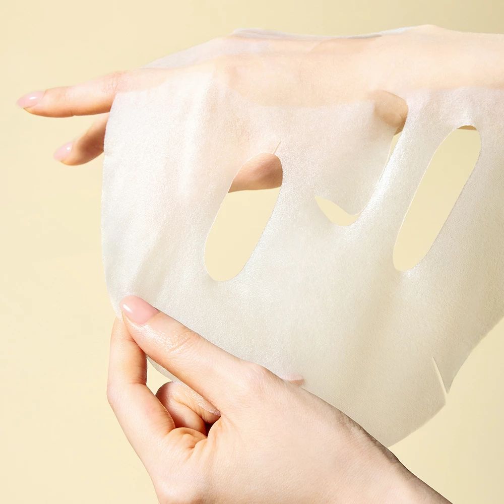 Hände halten eine weiße Gesichtsmaske mit Aussparungen für Augen und Mund.