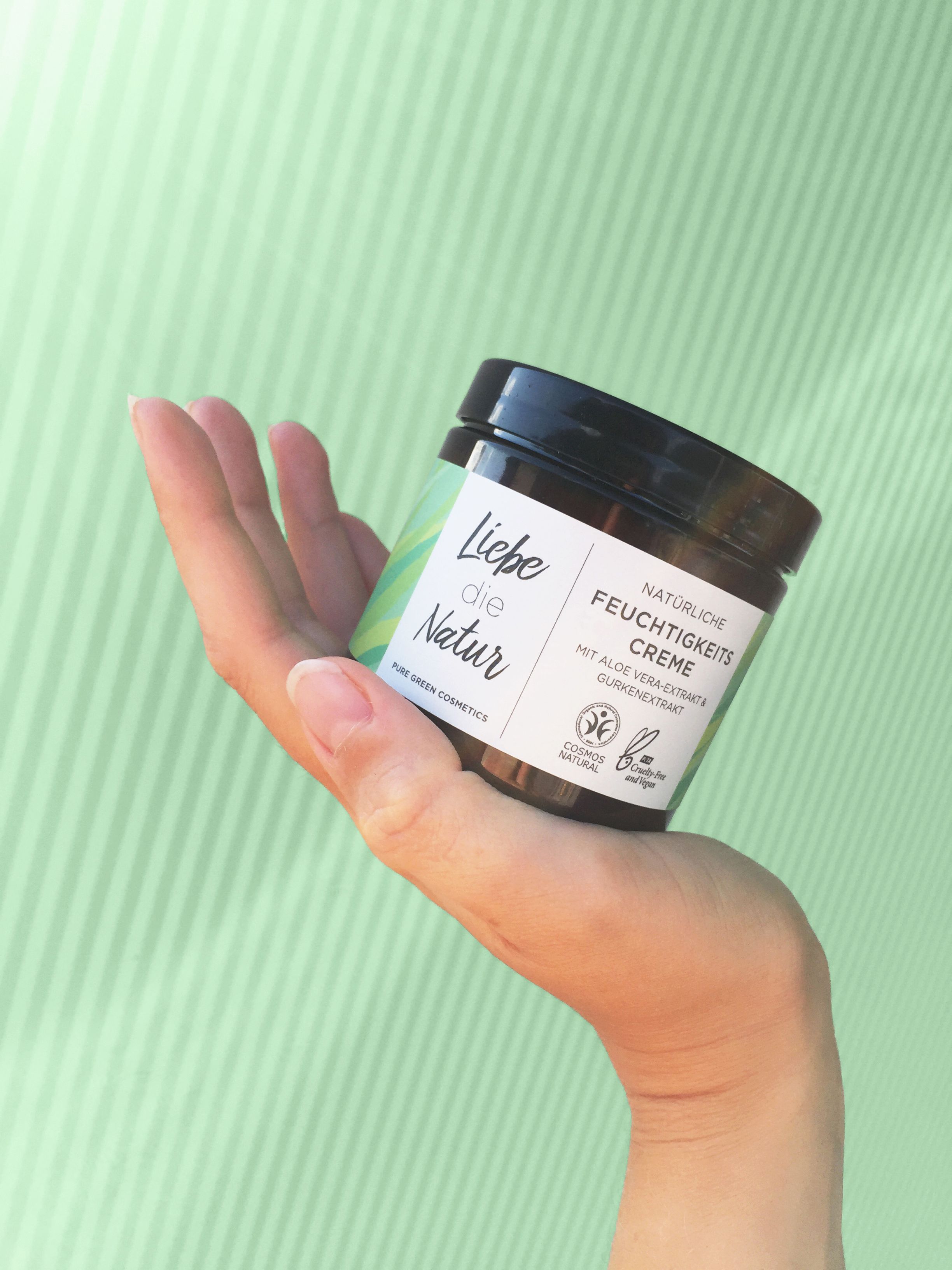 Dunkles Creme-Glas mit schwarzem Deckel, gehalten in einer Hand. Aufschrift: Liebe die Natur, Feuchtigkeitscreme. Grün-weißes Etikett.