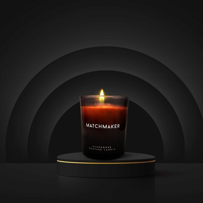Brennende Kerze auf einem Podest. Aufschrift: MATCHMAKER, PHEROMONE MASSAGE CANDLE. Schwarzer Hintergrund.