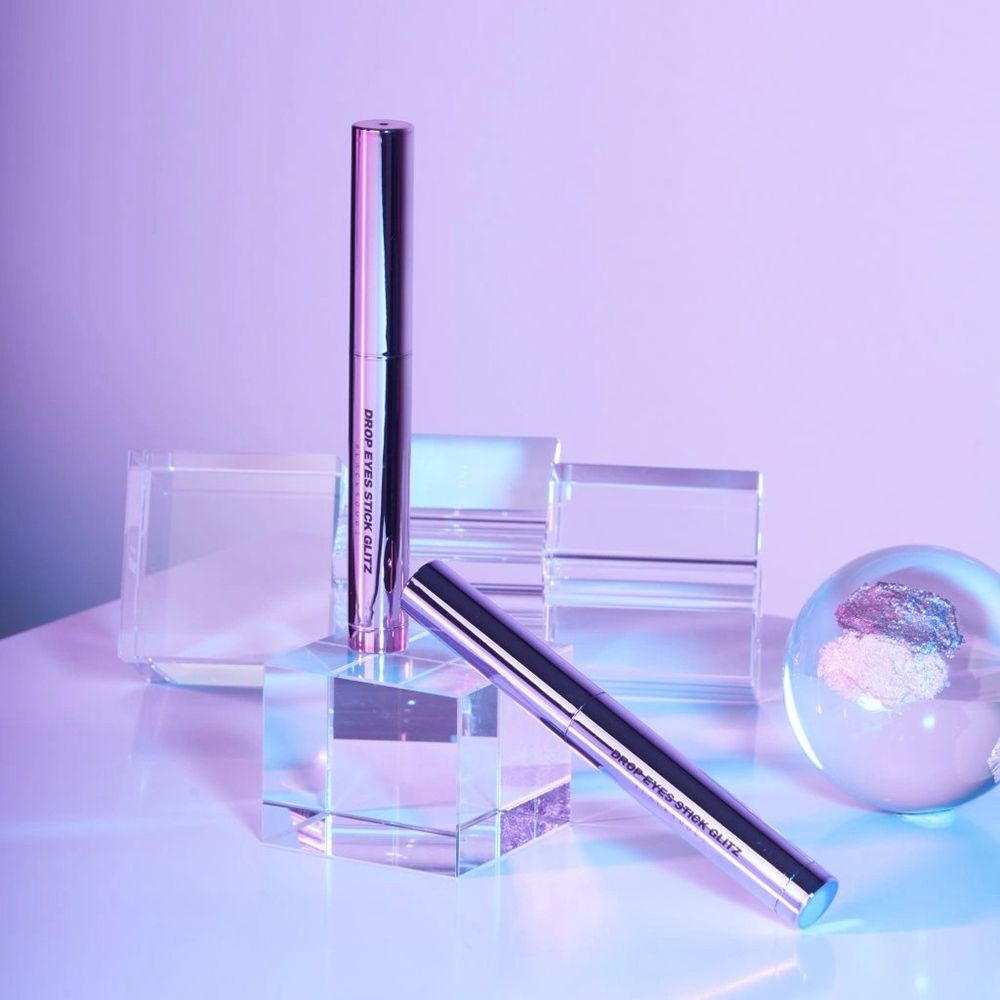 Zwei Drop Eyes Stick Glitz Lidschatten auf Glasblöcken. Ein Stift steht, einer liegt. Hintergrund mit Glasobjekten und Farbverlauf.