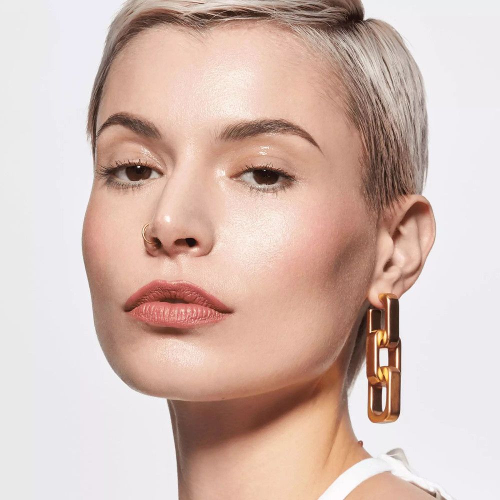 Porträt einer Frau mit kurzen blonden Haaren. Sie trägt rosafarbenen Lippenstift und goldene Ohrringe.