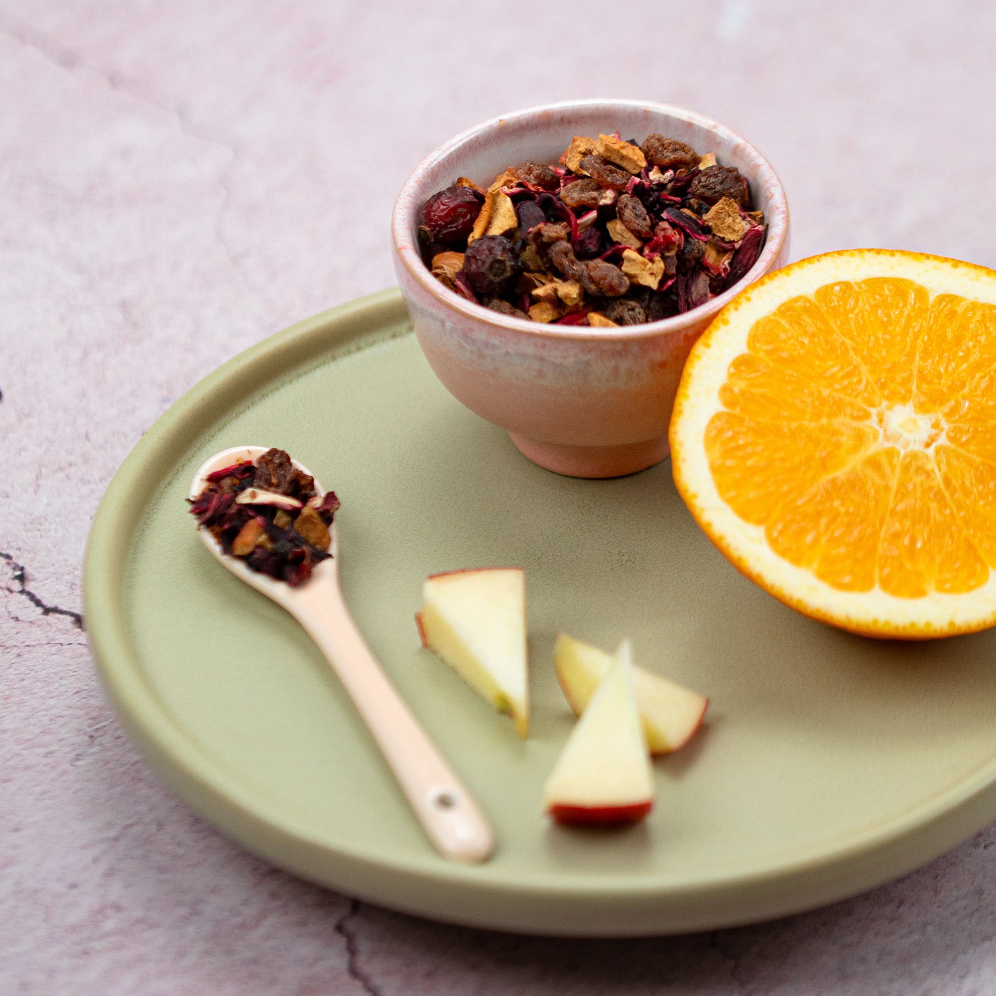 Schale mit losem Tee, daneben Löffel mit Tee und Apfelstücke. Halbe Orange.