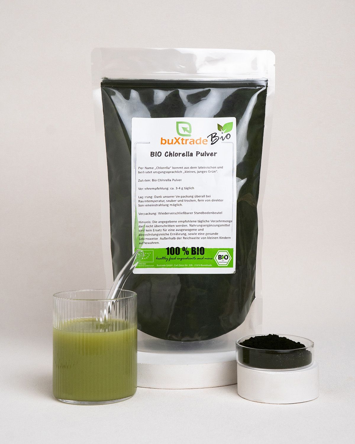 Glas mit grünem Getränk, daneben ein Behälter mit Chlorella-Pulver und ein Beutel mit Pulver. Auf weißem Podest.