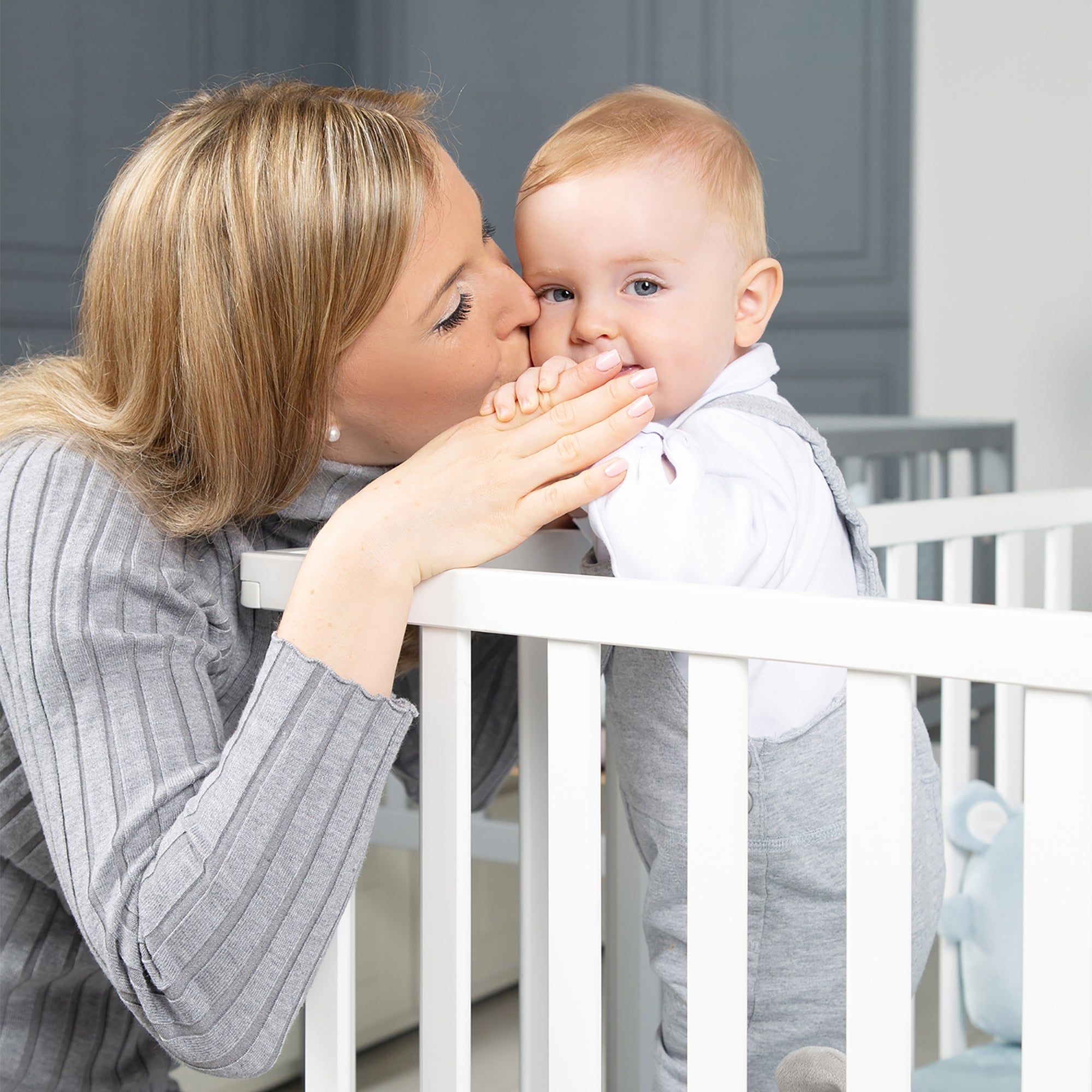 Mutter küsst Baby im Laufgitter. Weißes Holzgestell. Baby trägt Strampler. Grauer Hintergrund.