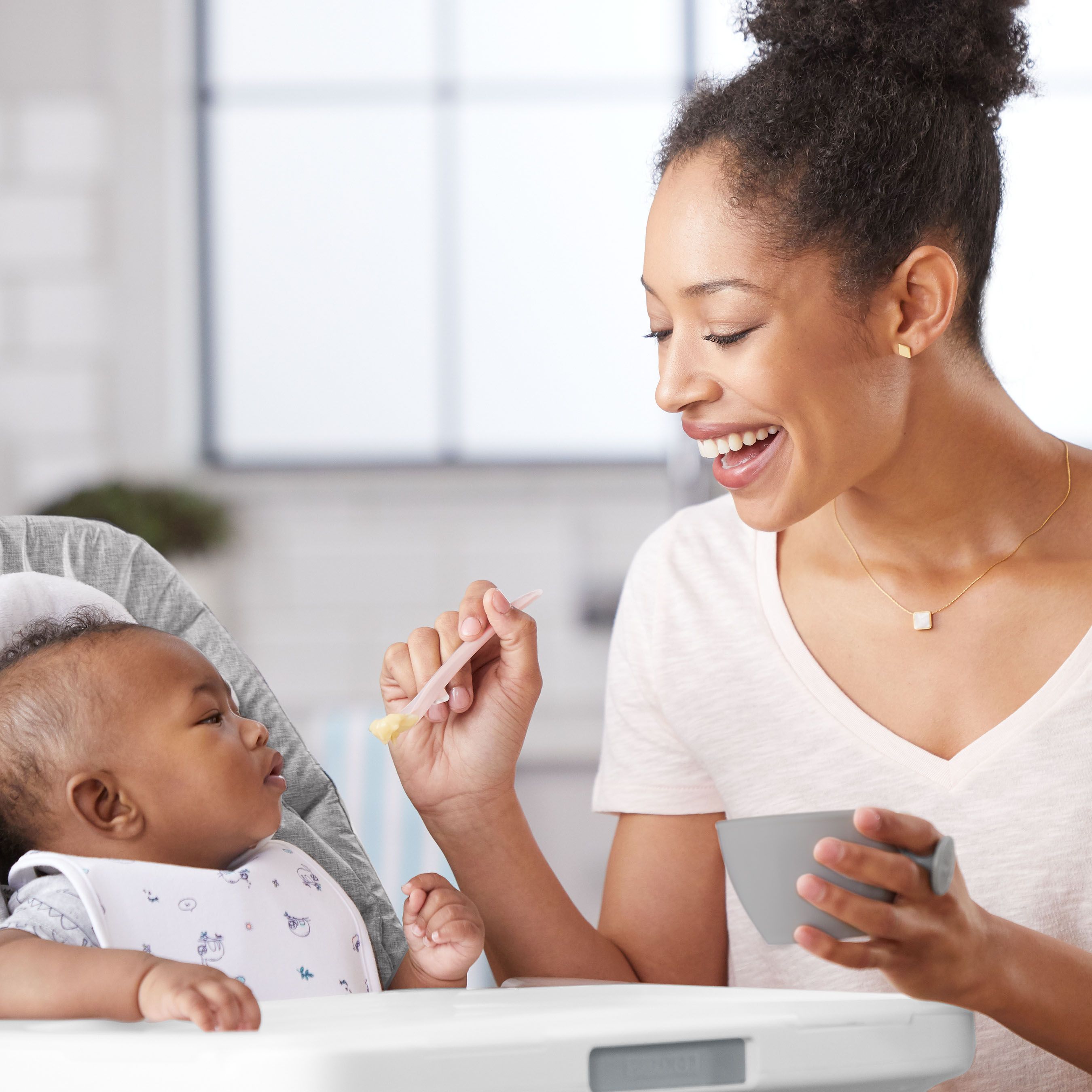 Eine Frau füttert ein Baby mit einem Löffel. Das Baby sitzt in einem Hochstuhl. Die Frau hält eine Schüssel.