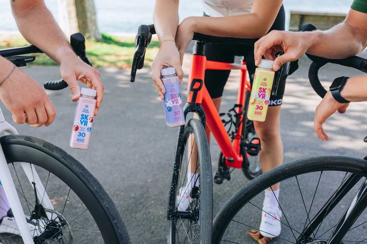 Drei Personen halten LiveFresh Skyr Protein Drink Flaschen. Sorten: Kirsche, Heidelbeere, Maracuja. Fahrräder im Hintergrund.