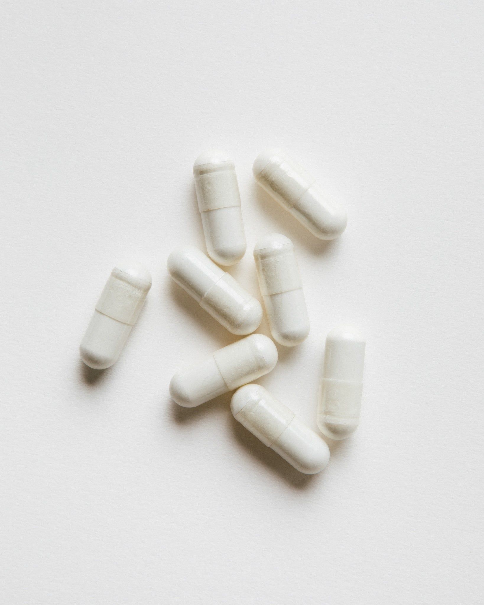 Acht weiße Kapseln liegen auf weißem Untergrund. Die Kapseln sind länglich und haben eine leicht abgerundete Form.