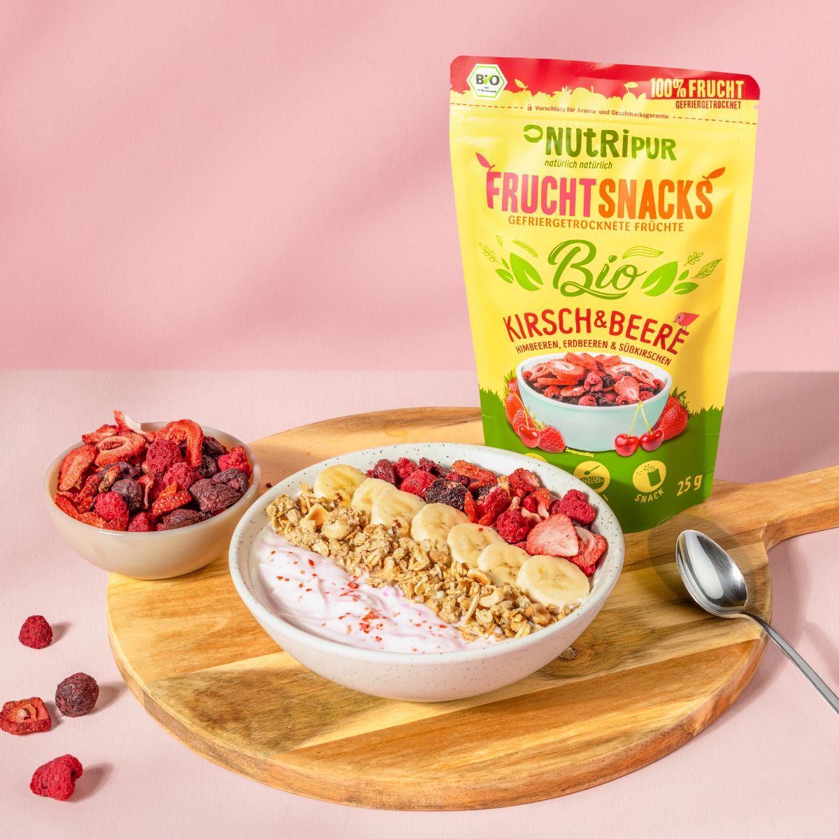 Gelbe Tüte "NutriPur Fruchtsnacks Bio Kirsch & Beere". Schale mit Joghurt, Müsli, Bananenscheiben und Früchten. Auf Holzbrett.