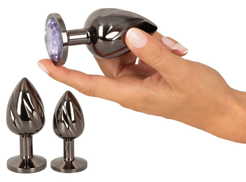 Hand hält einen Metall-Analplug mit lila Schmuckstein. Zwei weitere Plugs liegen daneben.