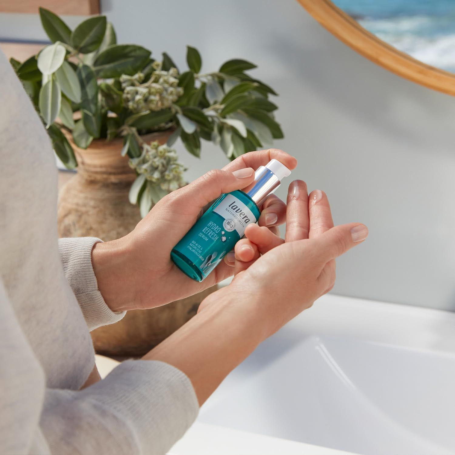 Person hält türkisfarbene Serumflasche. Aufschrift: lavera, Hydro Refresh Serum. Hände über Waschbecken. Hintergrund: Spiegel, Pflanzen.
