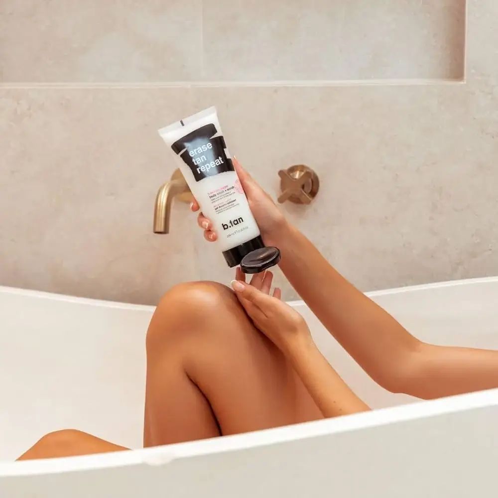 Person hält eine Tube über einem Bein in einer Badewanne. Auf der Tube steht "erase tan repeat" und "b.tan".