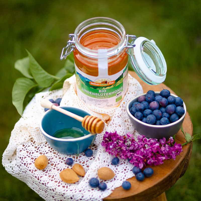 Honigglas "ImkerPur Bio Lindenblütenhonig" auf Holzschemel. Honig, Blaubeeren, Blüten und Honiglöffel.